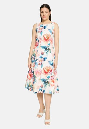 Frau trägt ein ärmelloses, knielanges weißes Kleid mit großem pinkem und blauem Blumenmuster, kombiniert mit weißen offenen High Heels.