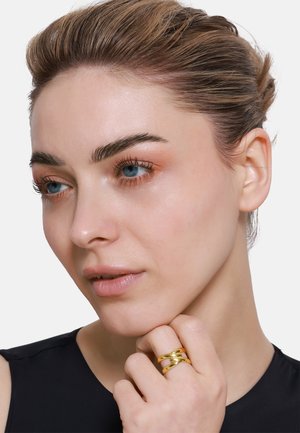 Gold metal ring with three curved bands, worn on fingers, showcasing a smooth, reflective texture against a dark top.