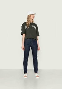 T-shirt oversized verde escuro com estampas gráficas, combinada com jeans azul escuro e sandálias brancas tipo slide. Um boné branco completa o visual.