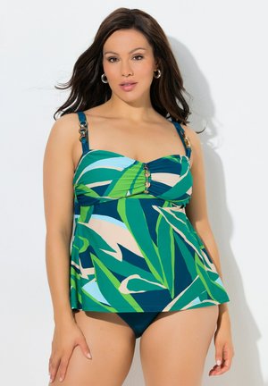 Kobieta ubrana w tankini w tropikalny wzór w zielono-niebieskich kolorach z detalami ze złotych guzików oraz pasujące majtki bikini, stojąca na białym tle.