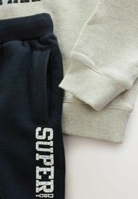 Sweater i lys grå med ribbede manchetter, parret med marineblå joggers med hvidt sprækket tekstdetalje. Begge genstande har en blød tekstur.