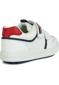 Sneaker bianco con tomaia in rete, accento rosso sul tallone e striscia laterale blu navy. Si chiude con una singola cinghia con velcro. Suola in gomma con grip testurizzato.
