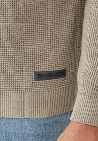 Beige Rippstrickpullover mit strukturiertem Strick, versehen mit einem schwarzen Etikett mit der Aufschrift "FÜR ALLE BEDINGUNGEN GEMACHT." Die Bündchen und der Saum sind ebenfalls gerippt.