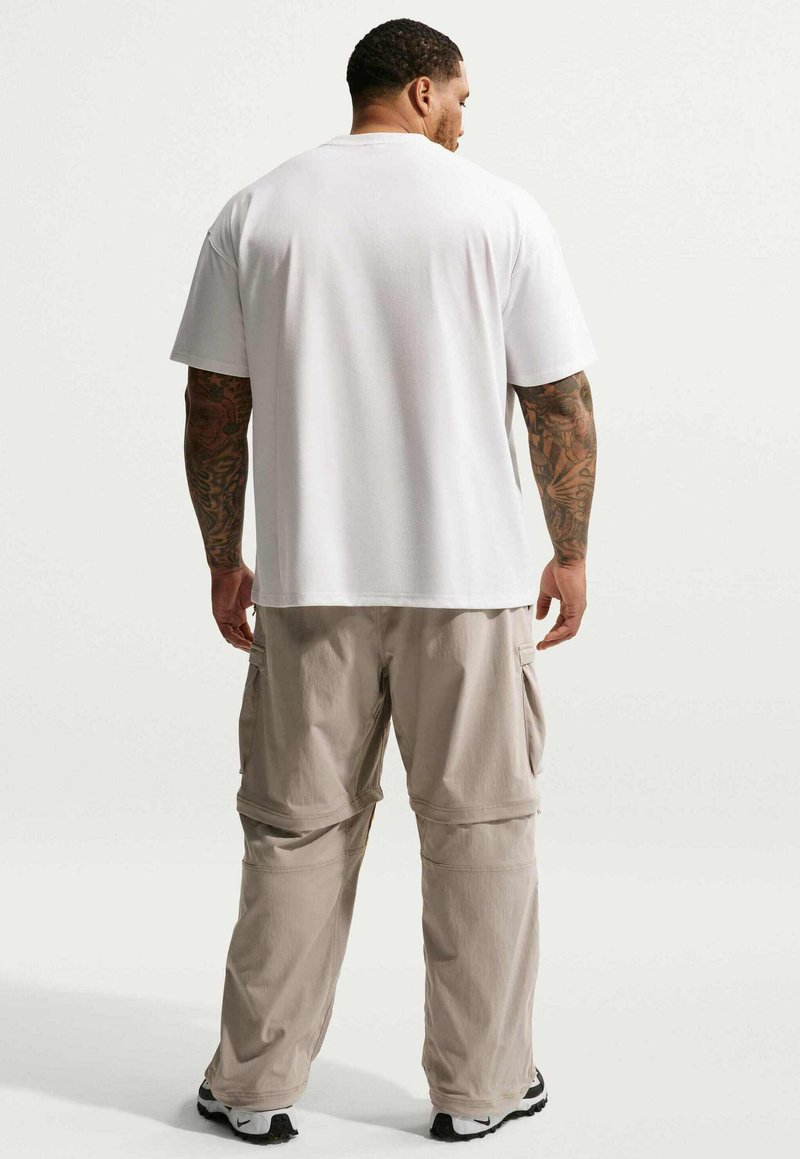 Homme aux bras tatoués vu de dos, portant un t-shirt blanc uni, un pantalon cargo beige et des baskets noir et blanc.