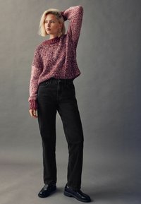 Maglione lavorato a maglia in tonalità di rosa e bianco, vestibilità rilassata con polsini a coste. Abbinato a jeans neri a vita alta e scarpe nere, il tutto su uno sfondo grigio.
