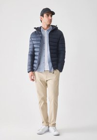 Navy puffer jacket with hood, zip closure, and quilted design, worn over a light blue shirt and beige pants, paired with white sneakers.