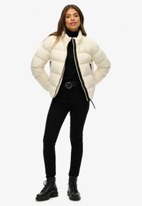 Hvid pufferjakke med høj krave og sort lynlås, båret over en sort turtleneck, parret med sorte skinny jeans og sorte støvler.