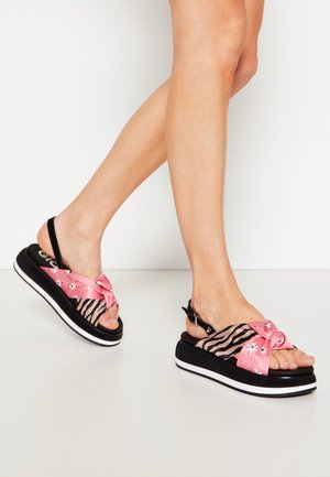 Personne portant des sandales compensées noires avec des brides en tissu à motif floral rose et zèbre ainsi que des brides noires à la cheville sur un sol blanc.