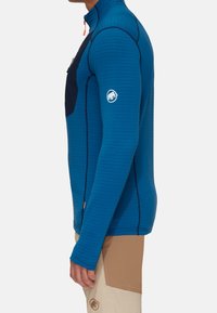 Chemise à manches longues bleue avec un col montant, motif texturé, poche poitrine noire et logo sur la manche. Présente des détails de couture contrastants.