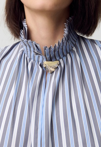 Blusa a rayas con rayas verticales azules y grises, un cuello con volantes y un cierre dorado en forma de corazón en el cuello.