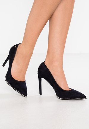 Sorte ruskind højhæle med spids tå og stiletthæl. Udstyret med sølvfarvede nitter langs den forreste kant. Glat tekstur.