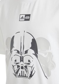 Bílý bavlněný tričko se černou grafikou tváře Darth Vadera, s logem Adidas nahoře a designem stormtroopera v pozadí.