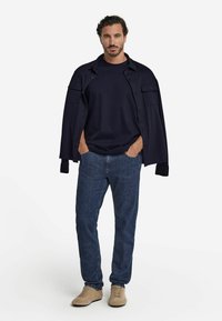 Mörkblå bomullsskjorta i jackstil över en mörkblå t-shirt med rund hals, kombinerad med blå denimbyxor och ljusbeige skor. Enkel design utan mönster.