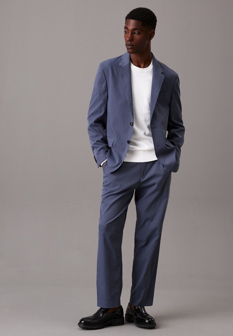 Uomo con un abito blu, un maglione bianco e mocassini neri, in piedi con le mani in tasca e che guarda di lato su uno sfondo grigio.