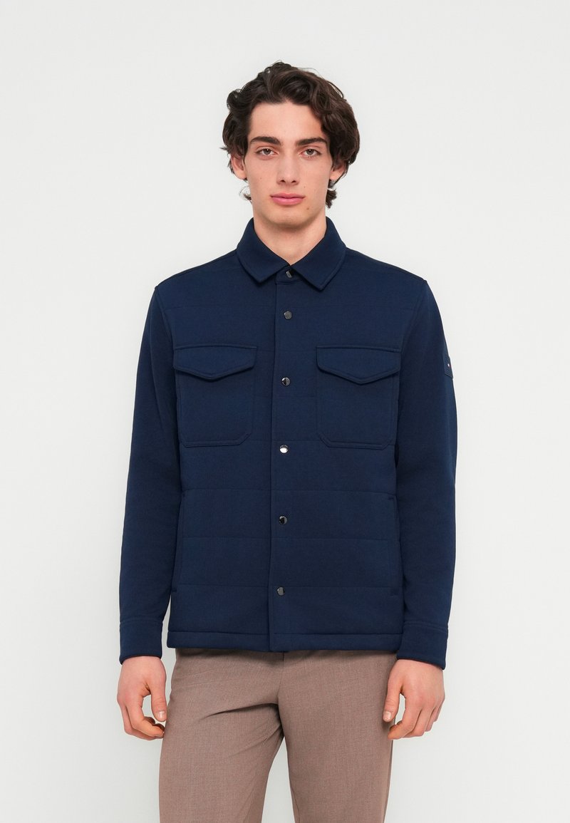 Jeune homme portant une veste bleu marine boutonnée avec des poches à rabat et un pantalon marron clair sur un fond blanc uni.
