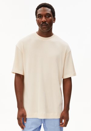 Homme aux cheveux courts bouclés et moustachu, portant une chemise ample crème à manches courtes et un pantalon bleu clair, debout sur fond blanc.