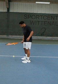 Joueur de tennis en t-shirt noir et shorts blancs, tenant une raquette de tennis orange, prêt à frapper une balle de tennis jaune sur un court bleu.