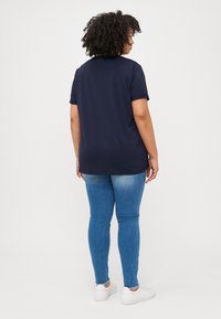 A person with curly hair wearing a navy t-shirt, blue jeans, and white sneakers stands with their back to the camera against a white background.