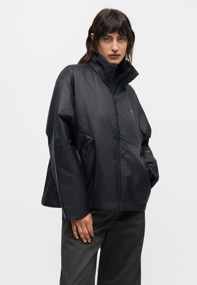 Άτομο που φοράει μαύρο φαρδύ μπουφάν με φερμουάρ και ψηλό γιακά και μαύρο παντελόνι, στέκεται μπροστά σε λευκό φόντο.