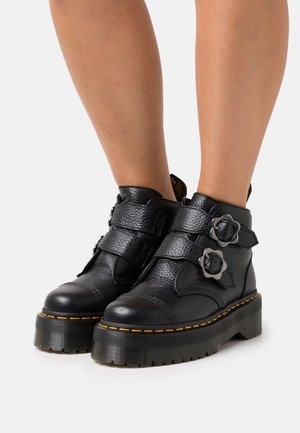 Schwarze Leder-Stiefeletten mit strukturiertem Finish, zwei verstellbaren Riemen, floralen Hardware-Akzenten, robusten Sohlen und kontrastierenden gelben Nähten.