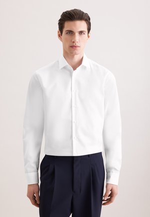 Homme aux cheveux foncés portant une chemise blanche à manches longues boutonnée et un pantalon plissé foncé, debout devant un fond clair uni.