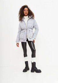 Giacca grigia imbottita con vita regolabile, cappuccio e chiusura con zip. Abbinata a leggings in finta pelle nera e stivali neri alti fino al ginocchio con lacci.
