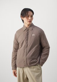 Brown lightweight jacket with a collared neck, visible logo patch, textured fabric, and button closure. Worn over beige trousers.
