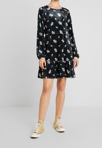 Svart sammetsklänning med långa ärmar, blommönster i rosa och grönt, utsvängd nederkant. Matchad med beige högskurna sneakers.