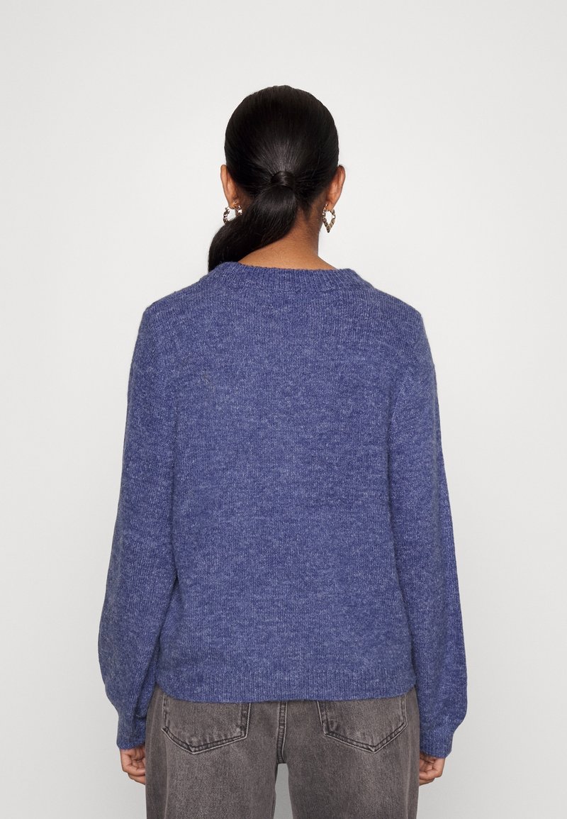 Blå stickad tröja med långa ärmar, rund halsringning och en texturerad yta. Bärs tillsammans med grå högmidjade jeans.