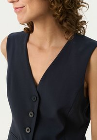 Marineblaues ärmelloses Kleid mit tiefem V-Ausschnitt, ausgestattet mit drei schwarzen Knöpfen auf der Vorderseite und einer figurbetonten Silhouette, glatter Stofftextur.