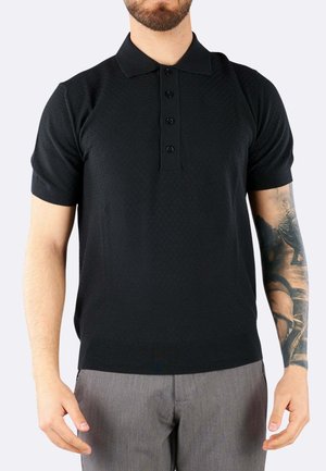 Uomo che indossa una polo nera a maniche corte con quattro bottoni e pantaloni grigi, che mostra un tatuaggio dettagliato sull'avambraccio destro.