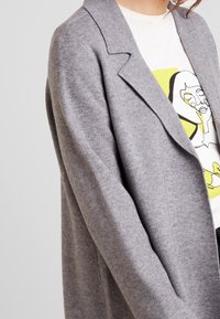 Šedý cardigan s měkkým, texturovaným povrchem, se zářezovým límcem a otevřeným předním dílem. Nesený přes bílou košili s uměleckým designem.