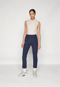 Polo sem mangas bege combinado com calças alfaiate azul-marinho escuras. Modelo usa ténis claros com detalhes em turquesa e meias brancas.