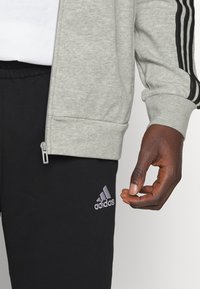 Siva zip-up jakna z črnimi črtami na rokavih, v kombinaciji s črnimi trening hlačami z odločeno sivo Adidasovo logotipom. Teksturirana tkanina, sproščen kroj.