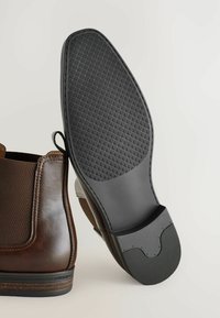 Acercamiento a una bota de cuero marrón con una suela negra texturizada, mostrando su diseño y construcción. El fondo es neutro.