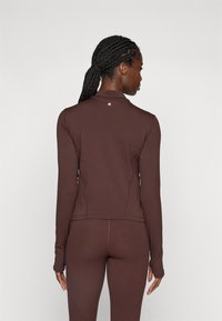 donna con capelli intrecciati che indossa un maglione aderente a maniche lunghe marrone e leggings abbinati, vista di spalle su uno sfondo neutro.