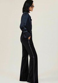 Vrouw in zwarte satijnen blouse en zwarte fluwelen broek met zijstreep, staande in profiel met handen in de zakken tegen een effen achtergrond.