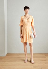 Robe à manches courtes et décolletée en V, de couleur jaune doux et à motif floral. Associée à des escarpins beiges à brides et un petit sac blanc. Fond en parquet bois.