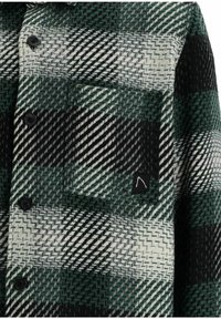 Knäppt rutigt skjorta med svarta, vita och gröna diagonala zigzag-mönster och en liten ficka med en svart etikett som har en vit logotyp.