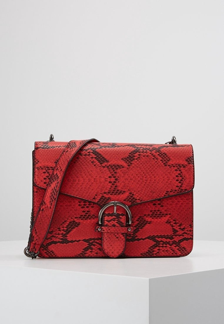 Punainen käärmeennahkakuvioinen käsilaukku, jossa on jämäkkä muoto, läpällä suljettava kansi ja pyöreä metallikoriste. Mukana säädettävä olkahihna.
