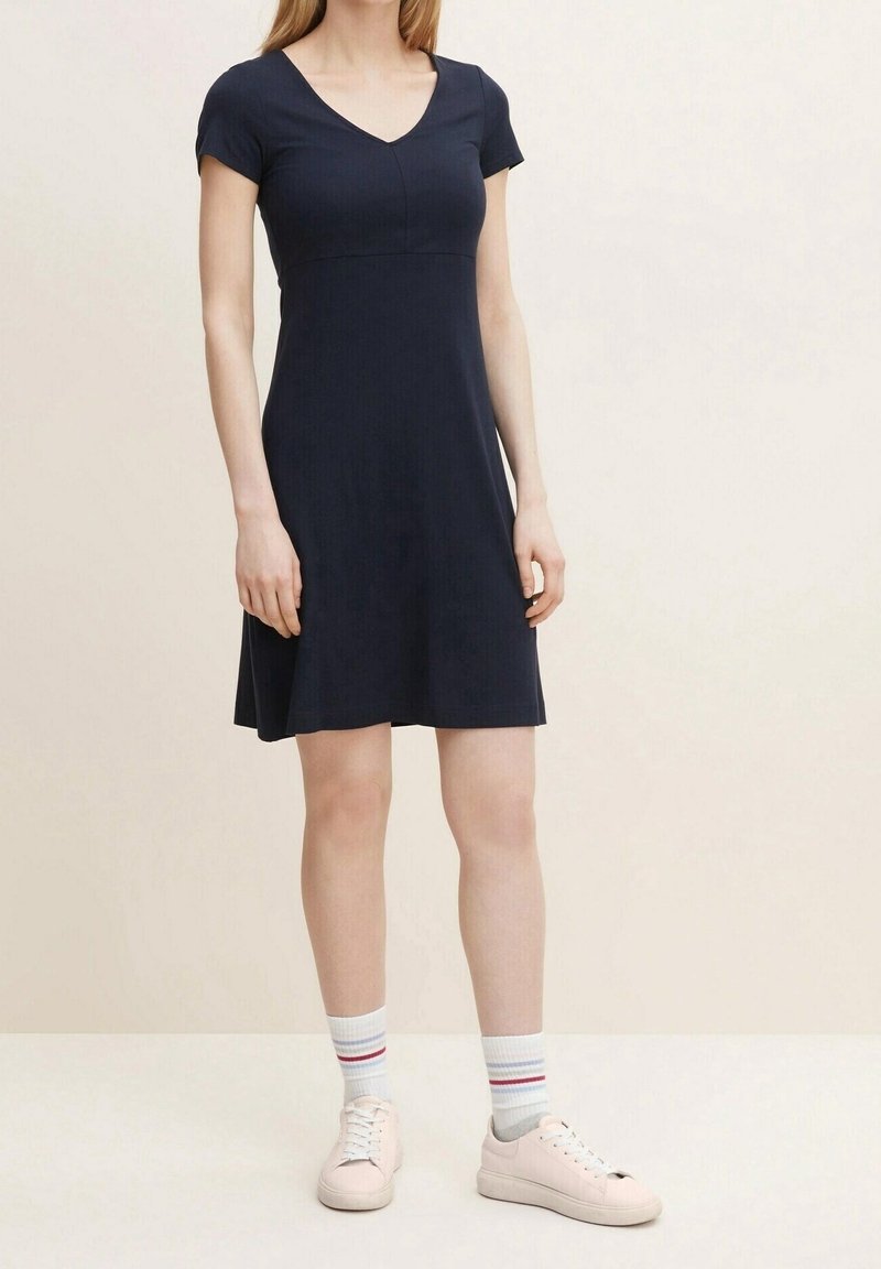 Mörkblå klänning med kort ärm och V-ringning, A-linjeformad silhuett, mjukt tyg, bärs med ljusrosa sneakers och randiga strumpor.