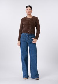 Pruun pehme kardigan, mille ees on nööbid, kombineeritud kõrge vöökohaga laia säärega siniste teksadega. Omab viit taskut ja elastset vöökohagi.