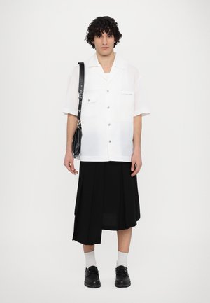 Camicia bianca a maniche corte con due tasche sul petto, gonna plissettata nera, scarpe nere e una borsa a tracolla nera. Colore uniforme, superficie liscia.