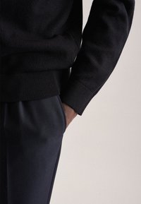 Maglione nero a maglia testurizzata con polsini a coste, abbinato a pantaloni scuri su misura. Mano posizionata nella tasca, mostrando un aspetto di tessuto liscio.