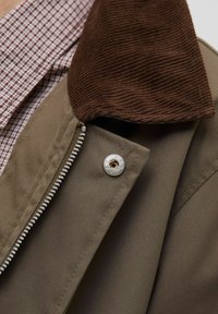 Bruine corduroy kraagjas met een tan lichaam, voorzien van een metalen rits en drukknoopdetails. Daaronder is een geblokt patroon shirt zichtbaar.