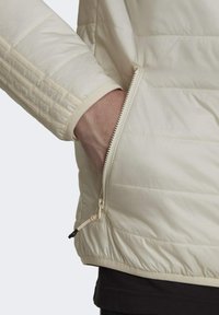 La giacca puffer di un crema chiaro presenta una tasca laterale con zip, polsini a costine e una finitura testurizzata, con cuciture verticali sottili per un tocco di dettaglio in più.