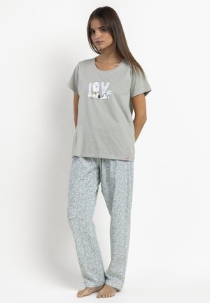 Joven mujer con camiseta gris clara con la palabra "LOVE" y un perro de dibujos animados, combinada con pantalones de pijama grises, sueltos y con estampado, de pie descalza.