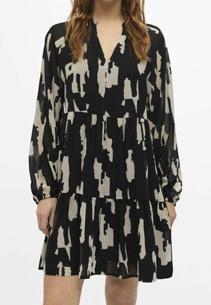 Woman wearing a black and beige abstract patterned long-sleeve dress with a V-neck and tiered skirt, standing against a plain background.