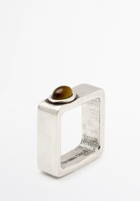 Quadratischer Silberring mit strukturiertem Oberfläche und einem braunen, polierten Stein, der oben in einer kreisförmigen Fassung eingelassen ist.