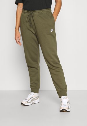 Person, die olivgrüne Nike-Sweathosen mit Kordelzug und weißen Turnschuhen trägt, vor einem schlichten hellen Hintergrund steht.